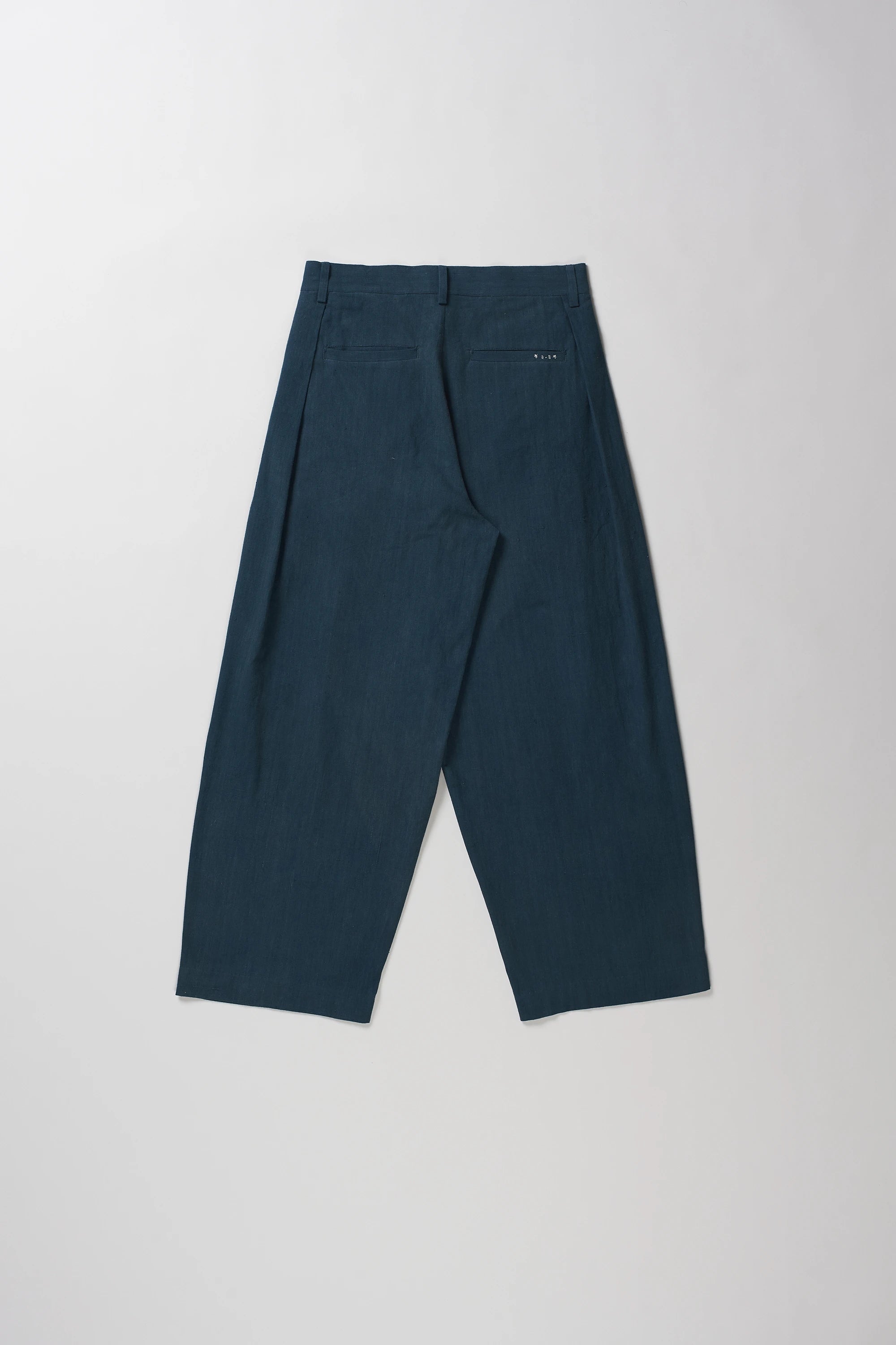 Back shot of pair of black indigo pleated trousers laid flat against a light gray background.