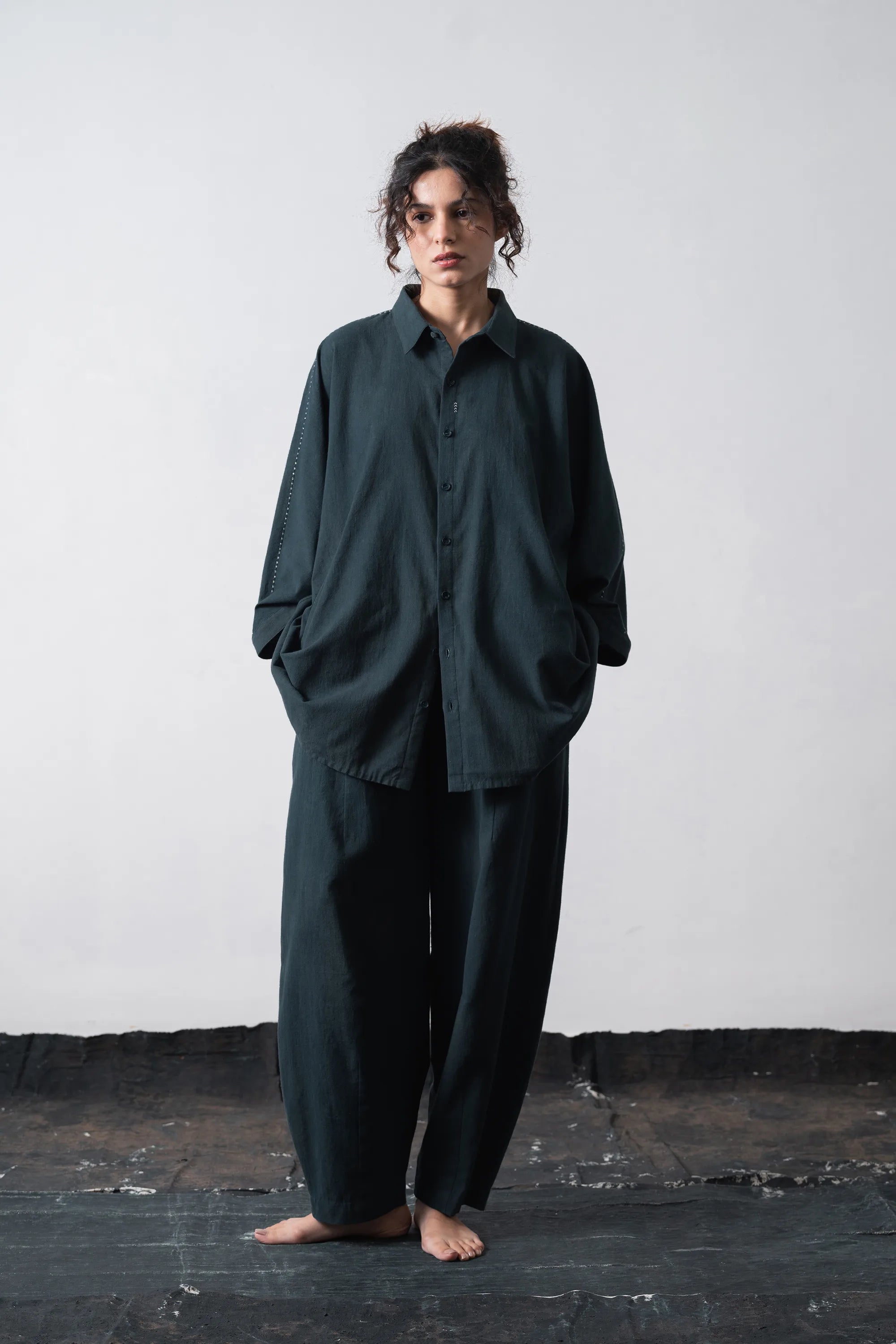 A model stands barefoot against a light grey wall and dark floor, wearing an oversized black indigo button-up shirt and matching wide-leg pants.