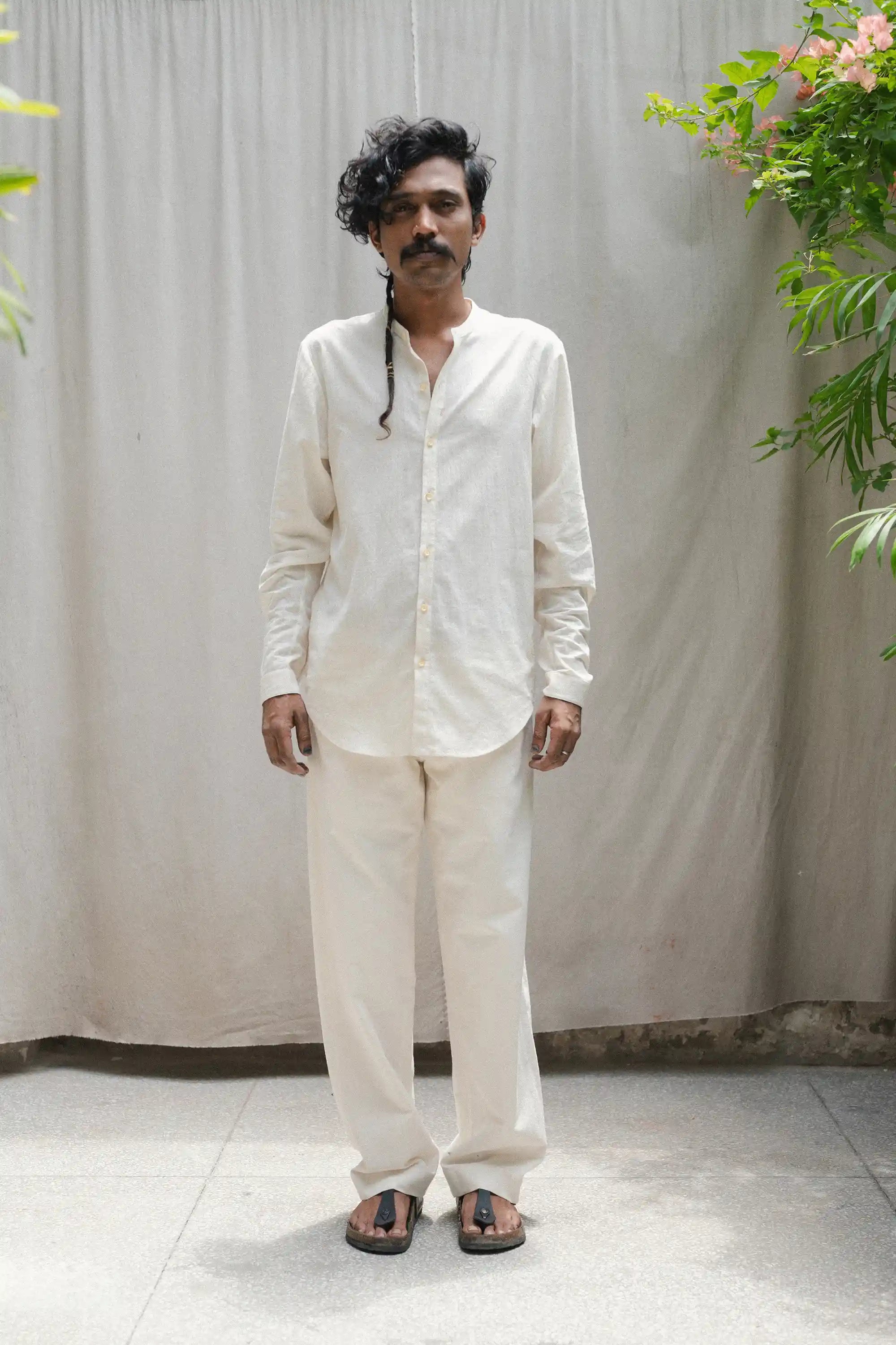 Man wearing a ecru shirt and pants standing against a plain background with plants on either side.