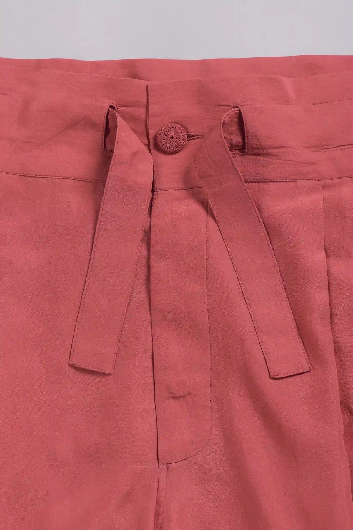 Close-up of a dusty rose pink garment, showing the waistband, button closure, and tie belt details.