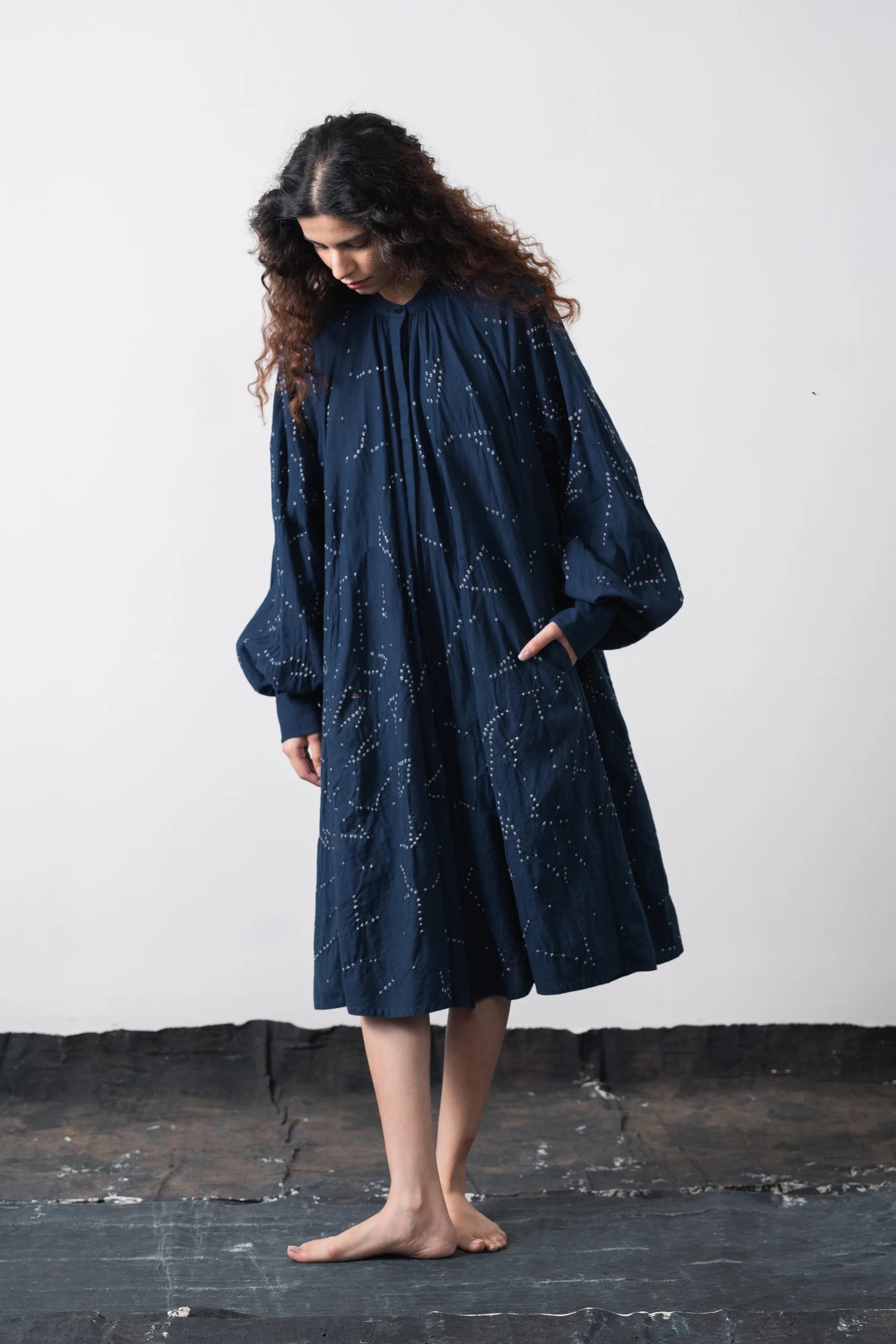A woman stands barefoot against a plain off-white wall, wearing a loose space blue dress with a bandhani artwork and voluminous long sleeves.