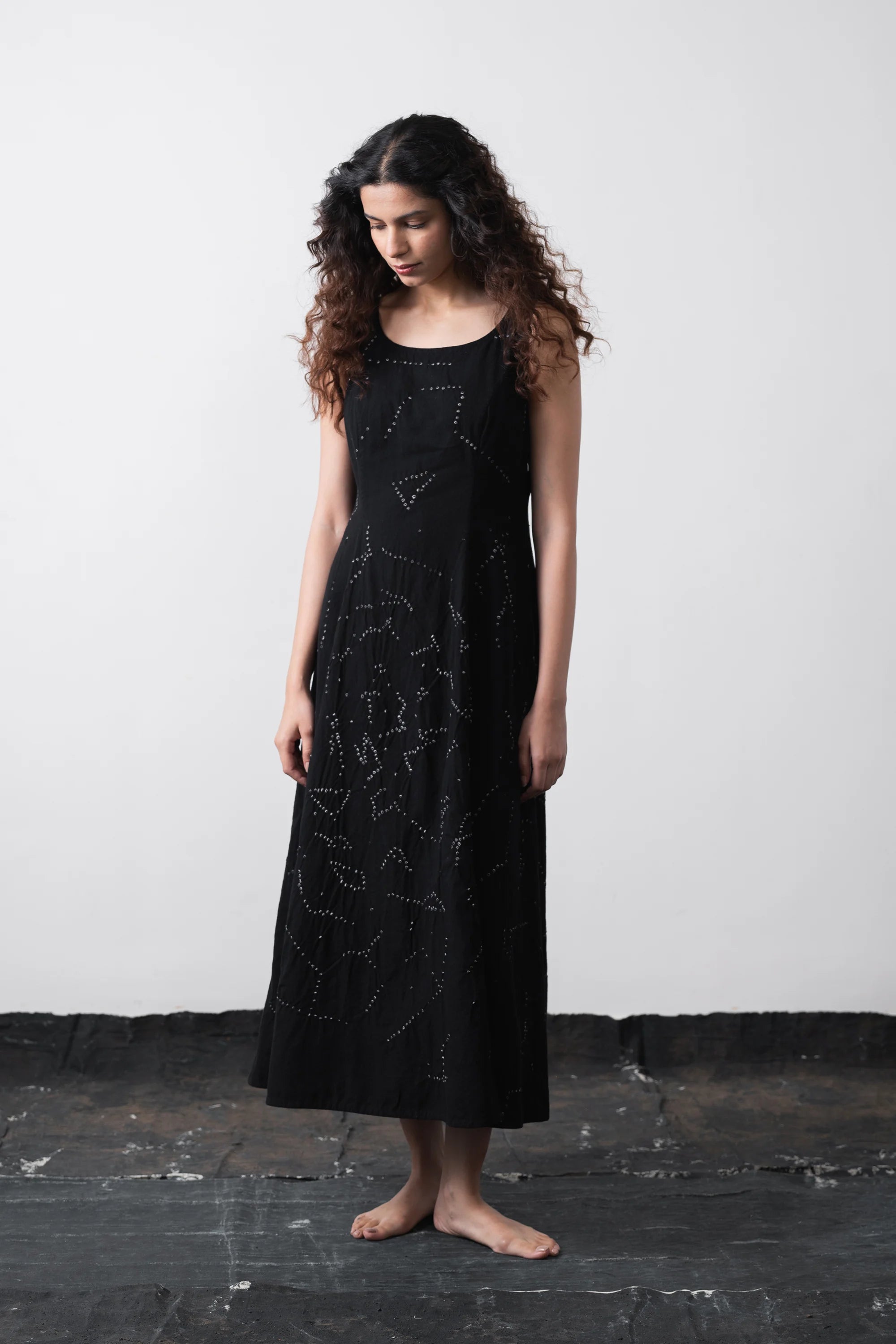 A woman stands barefoot against a plain light grey wall wearing a sleeveless black midi dress.