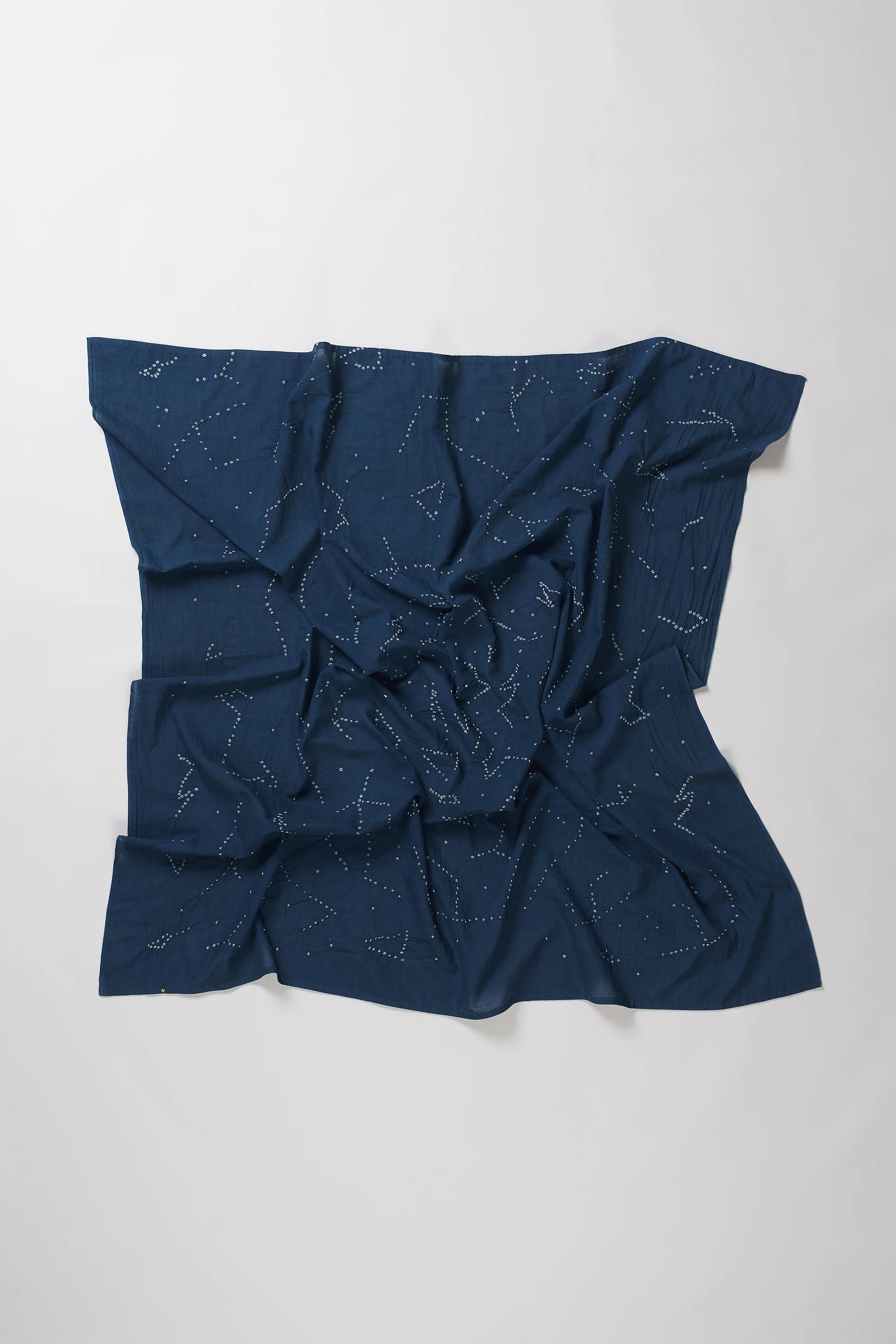 A space blue fabric accessory, likely a scarf or bandana, featuring a white celestial print of stars and constellations, laid out in a loose, crumpled square shape against a plain light grey background.