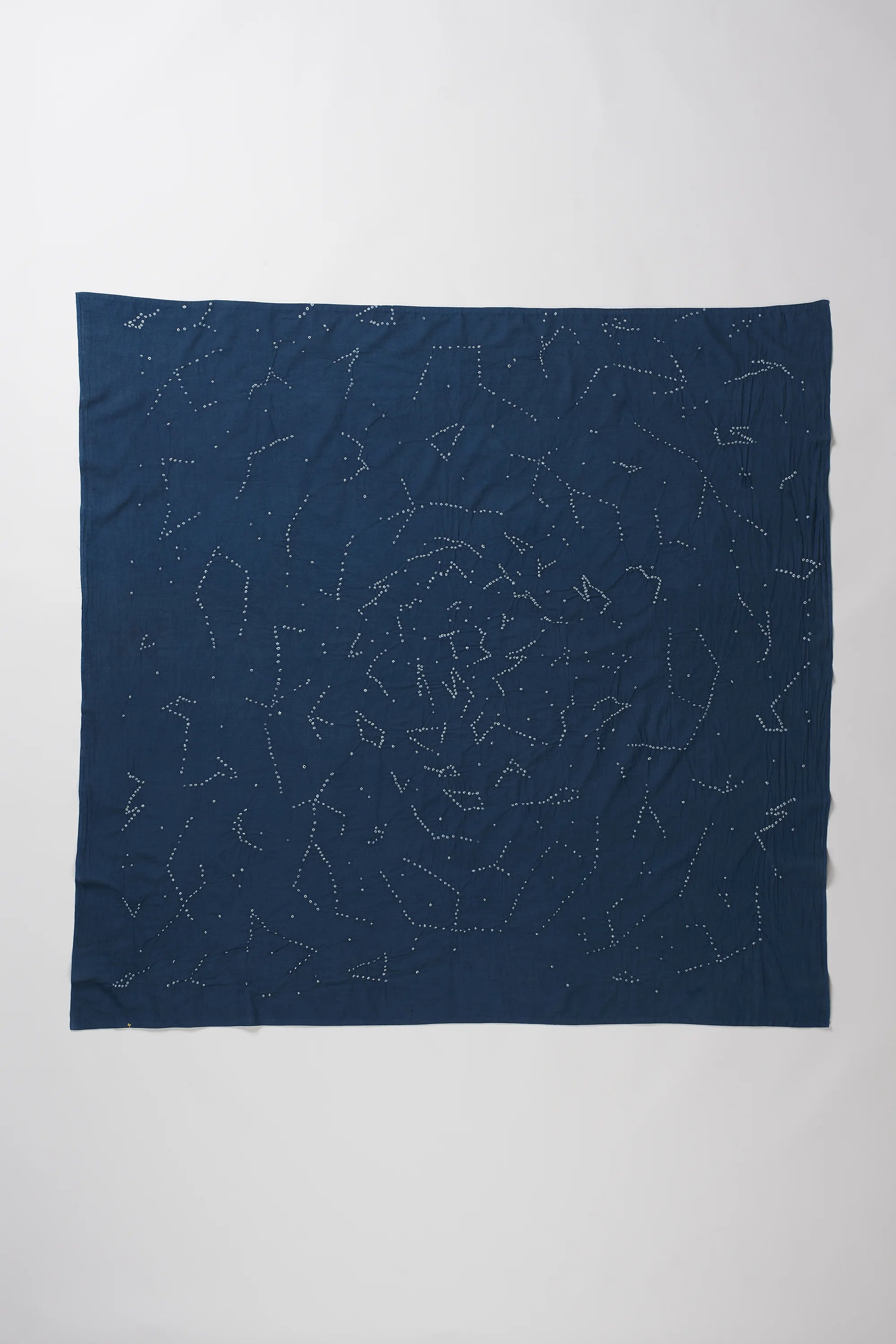 A space blue fabric accessory, likely a scarf or bandana, featuring a white celestial print of stars and constellations, laid out in a loose, crumpled square shape against a plain light grey background.