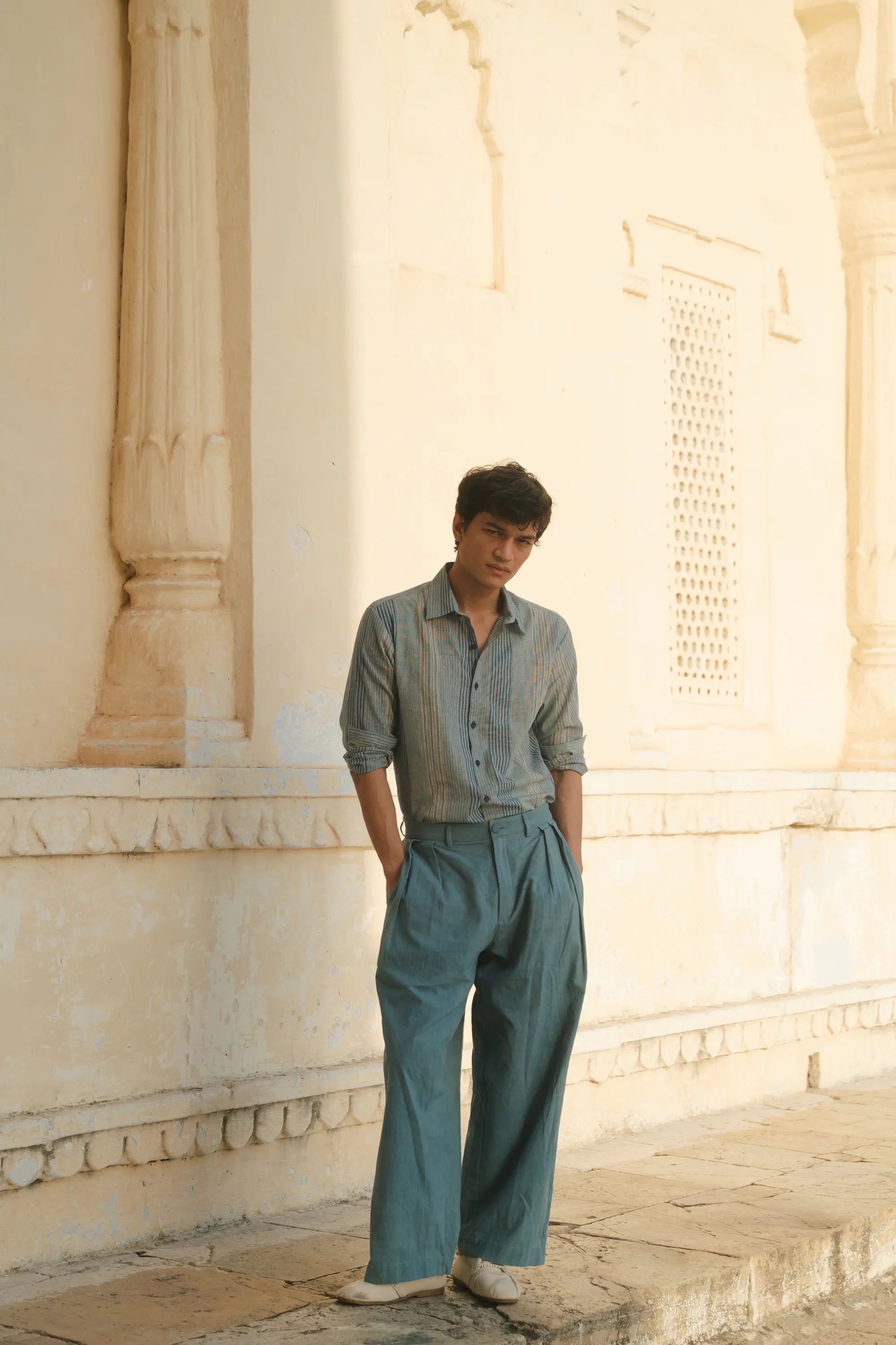 Man in a light blue shirt and wide-leg pants standing against a classical architecture background