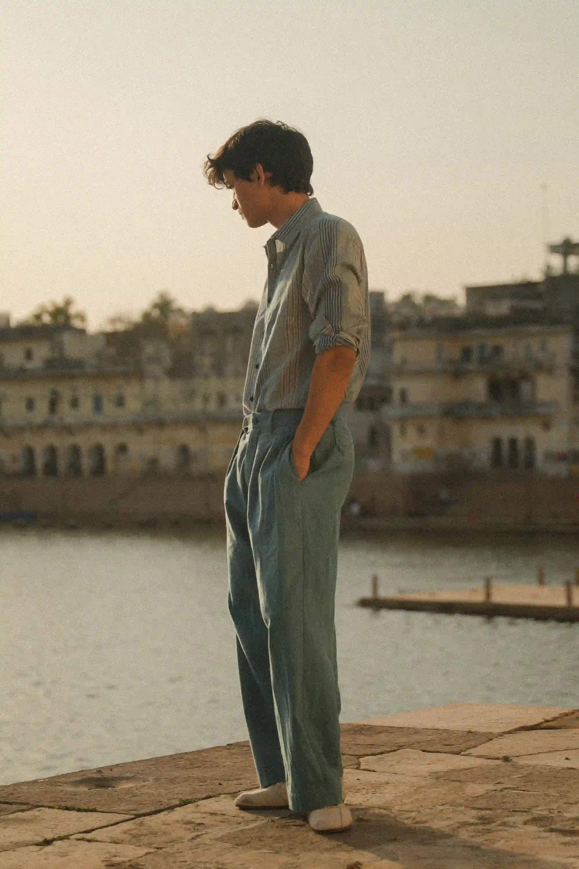 A side shot of a man wearing powder blue pant standing by a waterfront with buildings in the background