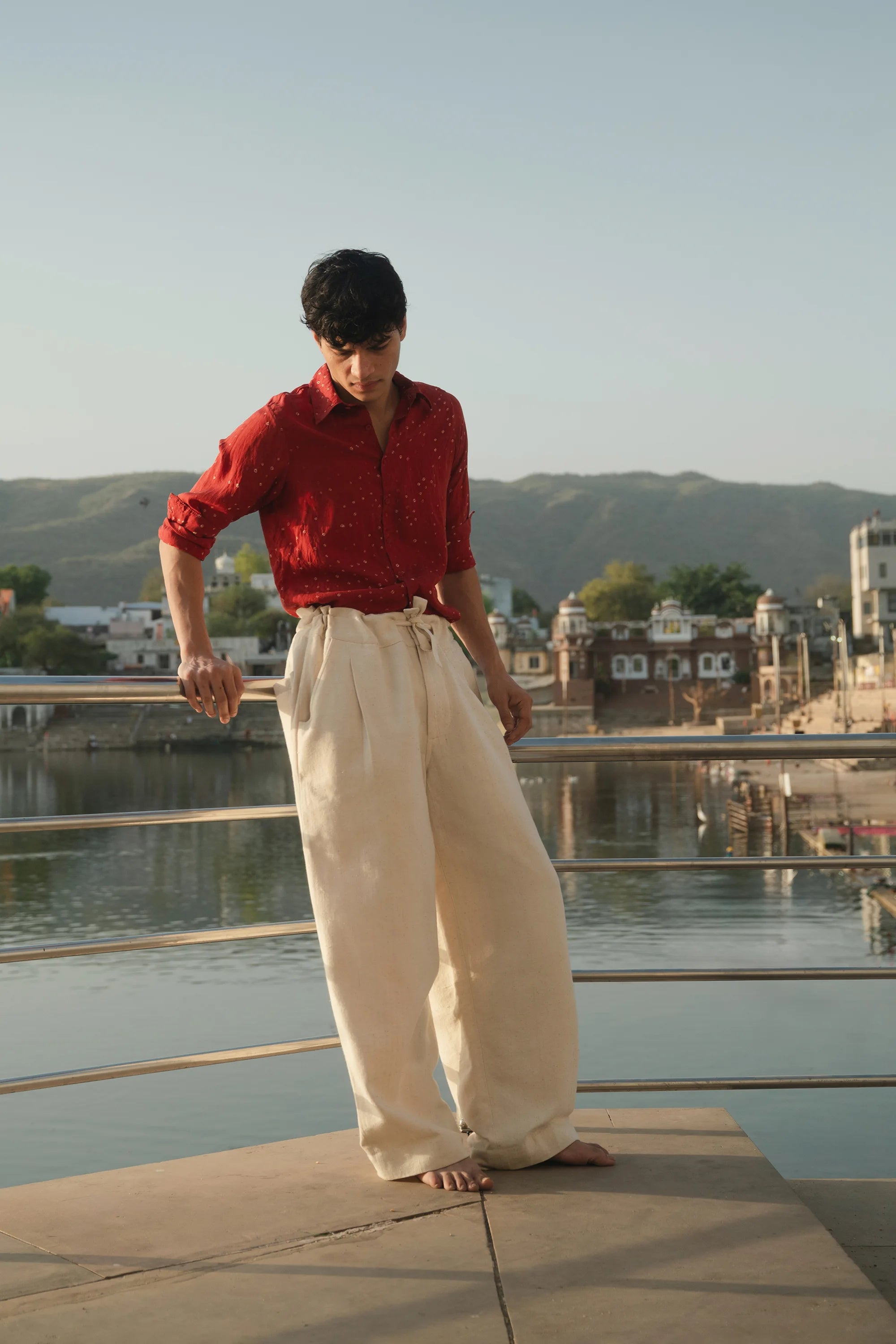 Person wearing a red shirt and white pants standing on a rooftop with a scenic view of mountains and buildings.