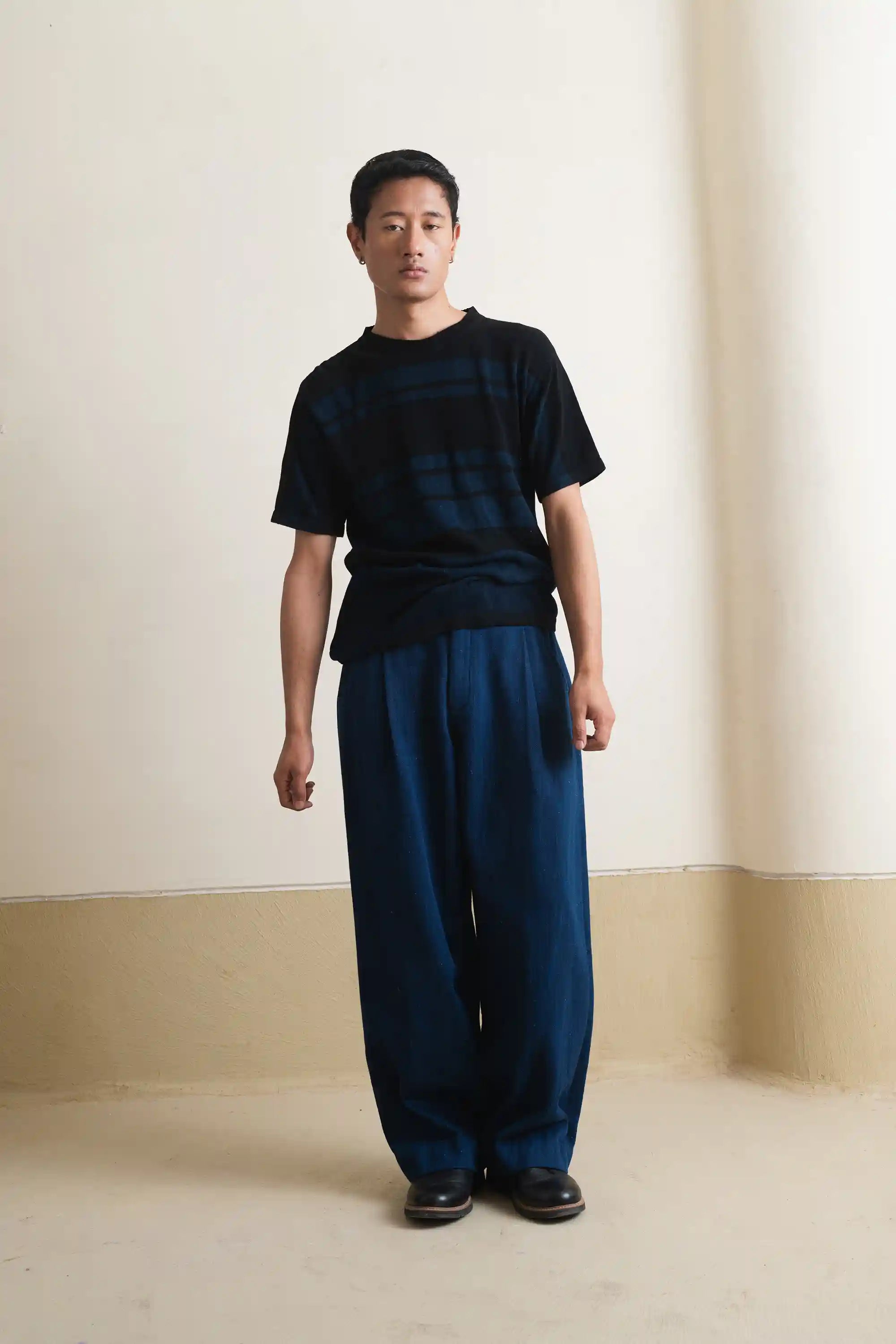 Man wearing a black t-shirt and blue pants standing against a plain wall.