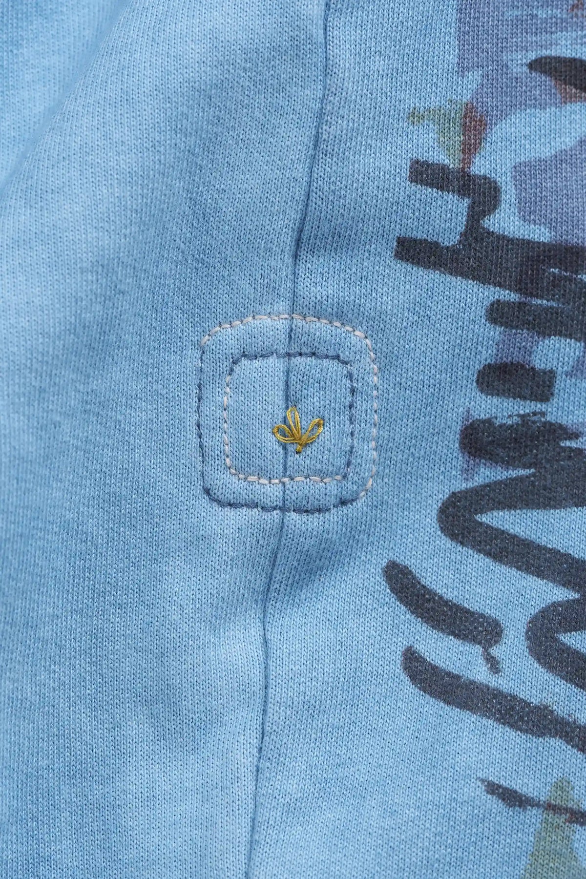 Close-up of a blue knit garment featuring a square patch with a small yellow embroidered leaf logo, alongside dark graphic lettering.