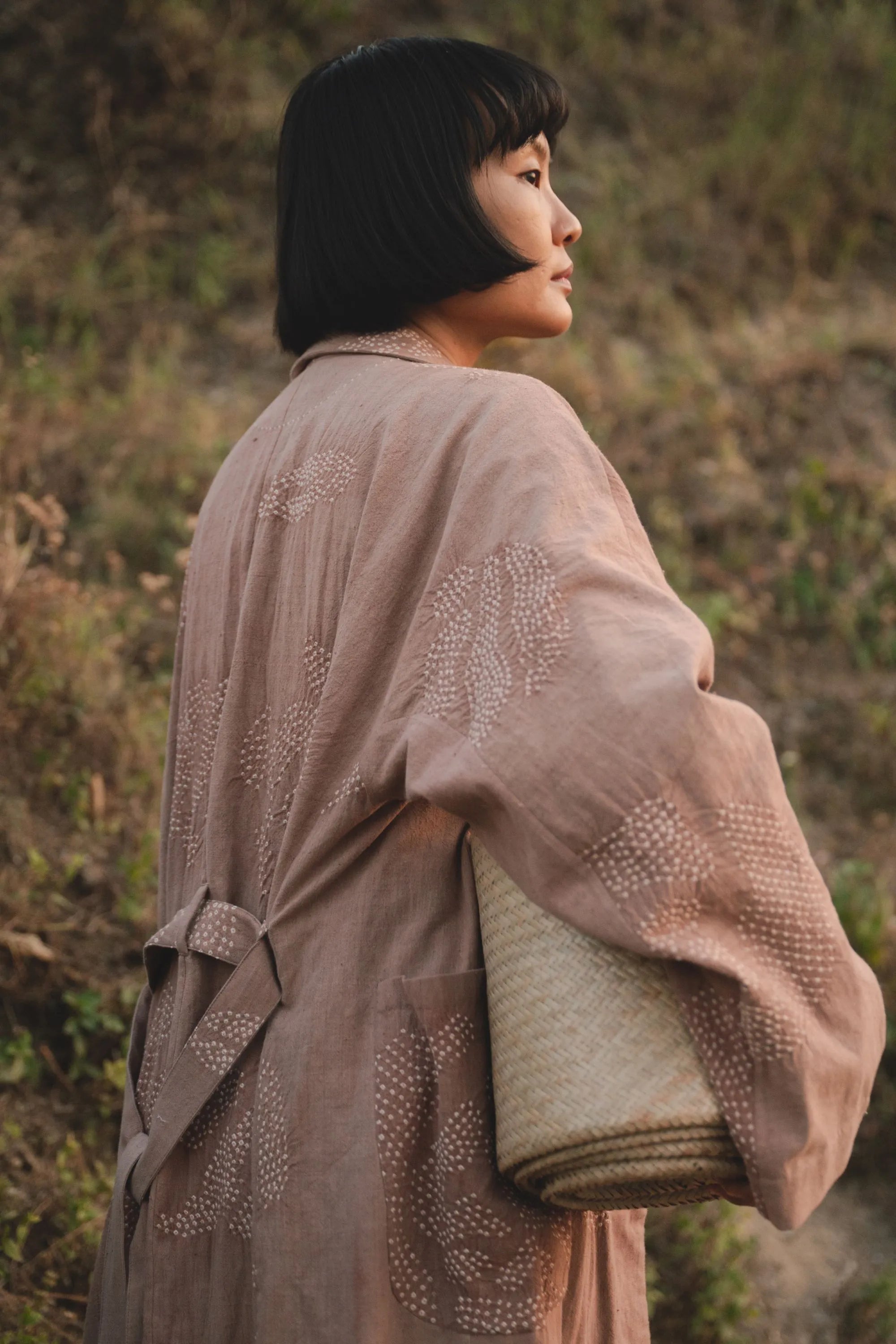 Woman in a light old rose cotton shrug holding a woven mat, standing in a natural setting.