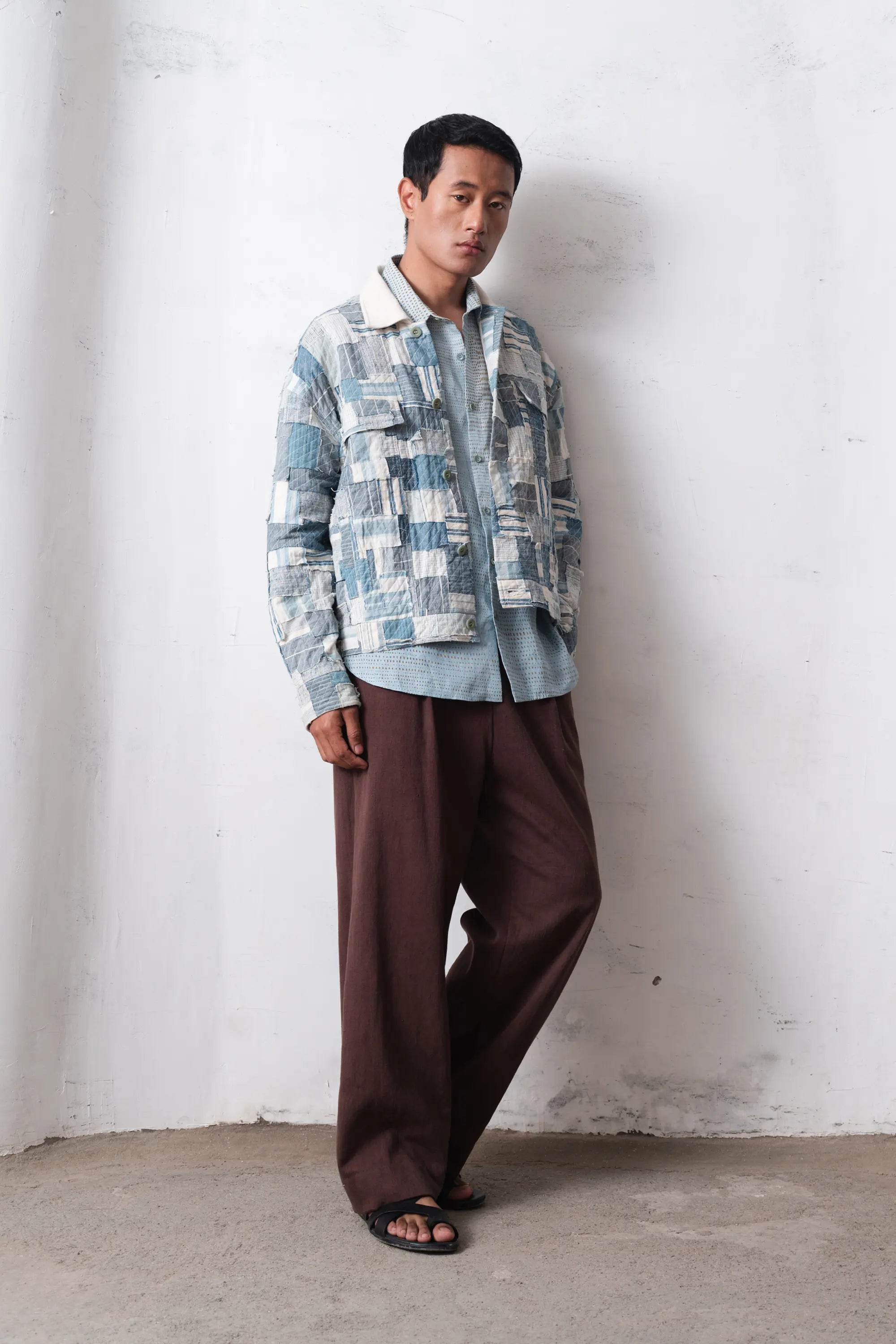 Man wearing a patchwork jacket and brown pants against a light grey wall