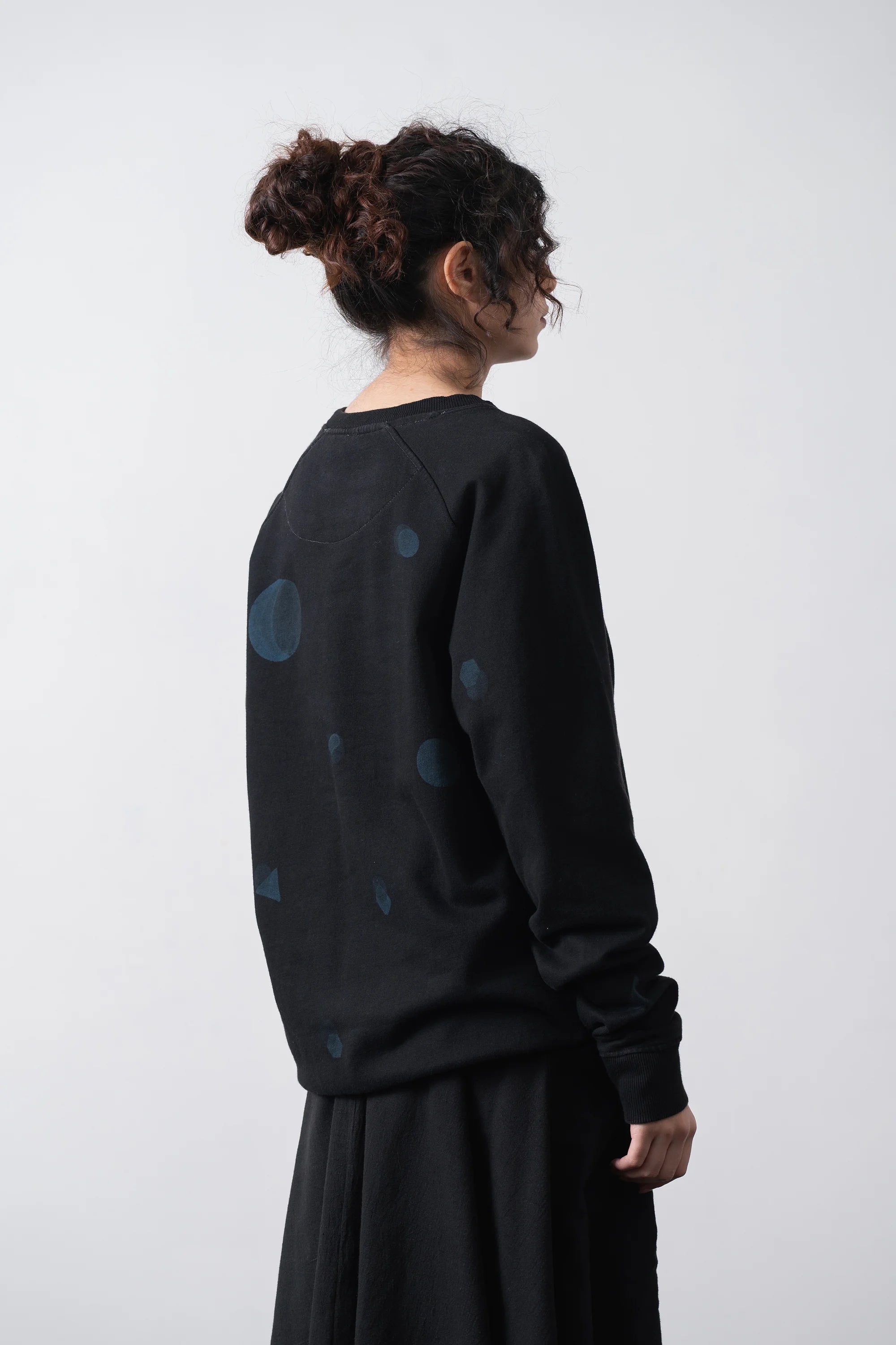A woman shown from the back wearing a black long-sleeve sweatshirt with scattered blue circular shibori, paired with a black skirt, against a plain light grey background.