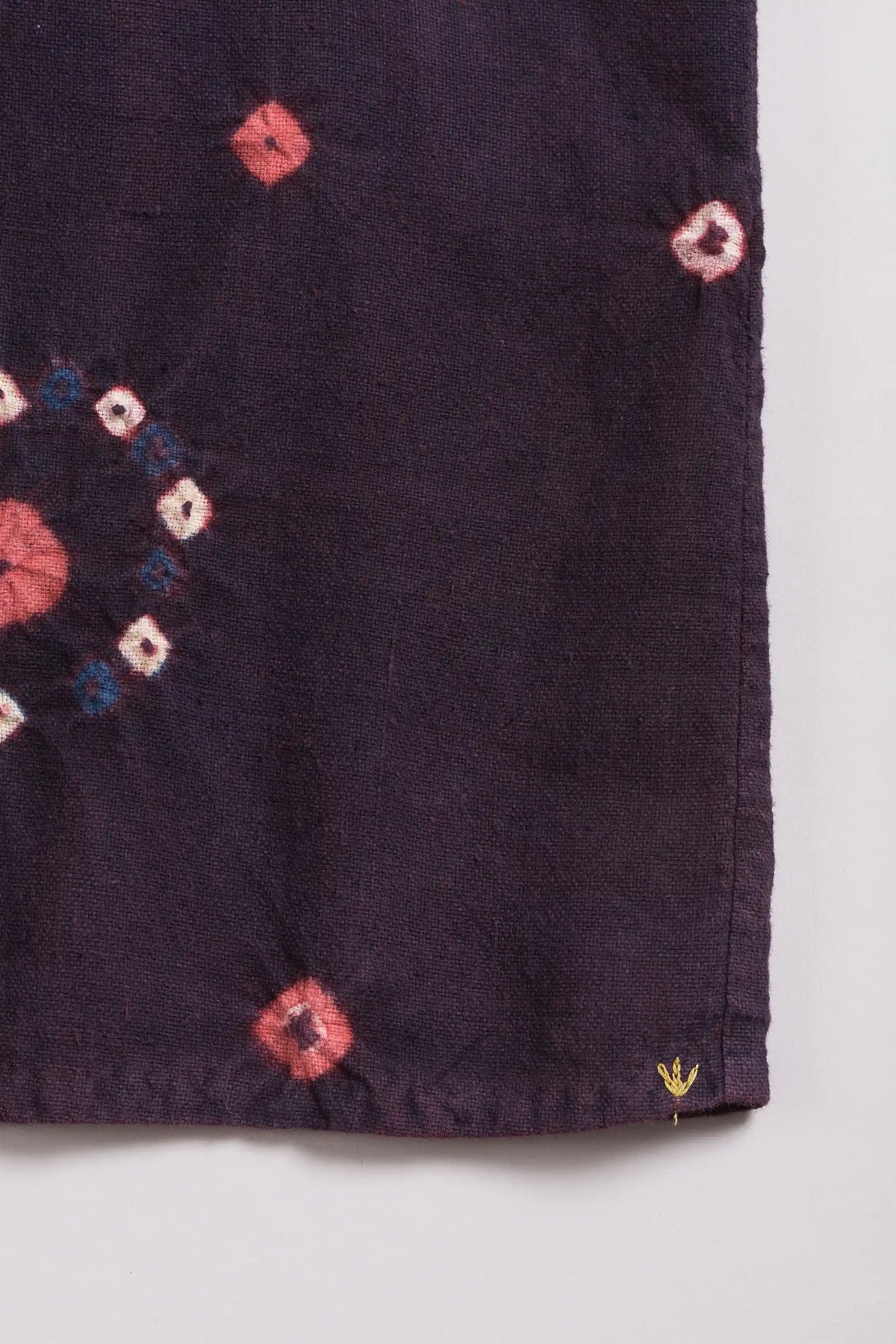 A close-up of a purple red fabric, likely cotton, featuring bandhani with a small yellow embroidered detail near the bottom right corner.