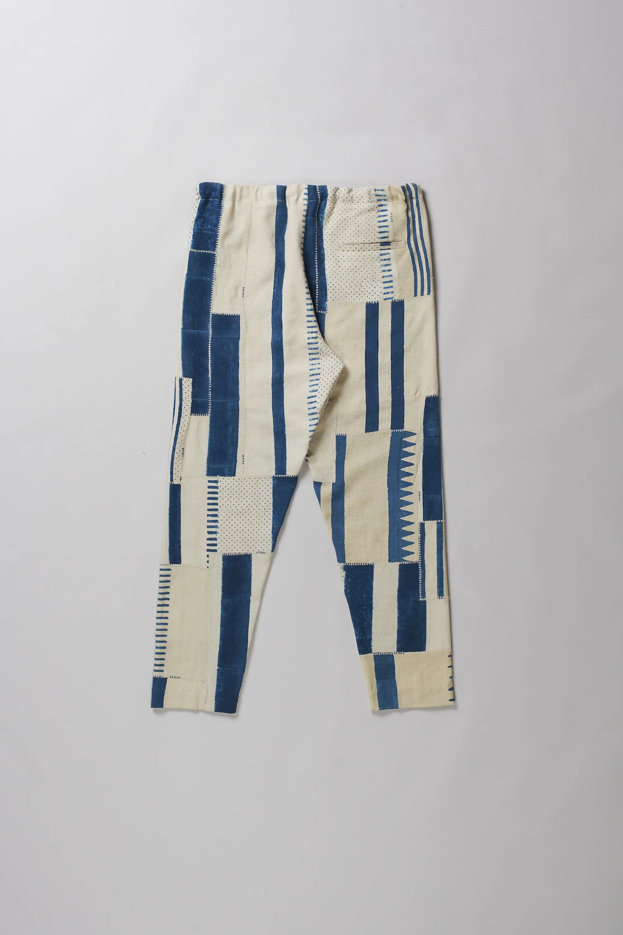 Back shot of pants with a geometric patchwork pattern in blue and cream, featuring a drawstring waist, laid flat on a light grey background.