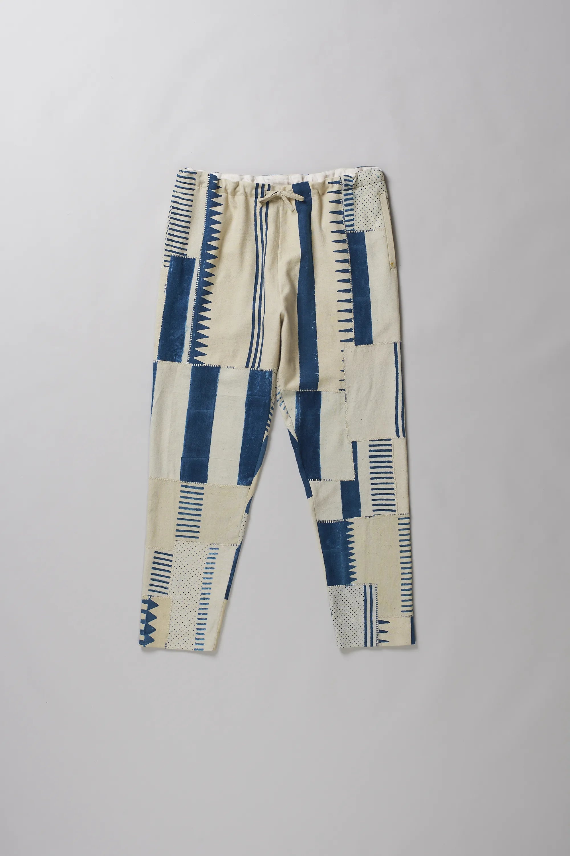A pants with a geometric patchwork pattern in blue and cream, featuring a drawstring waist, laid flat on a light grey background.